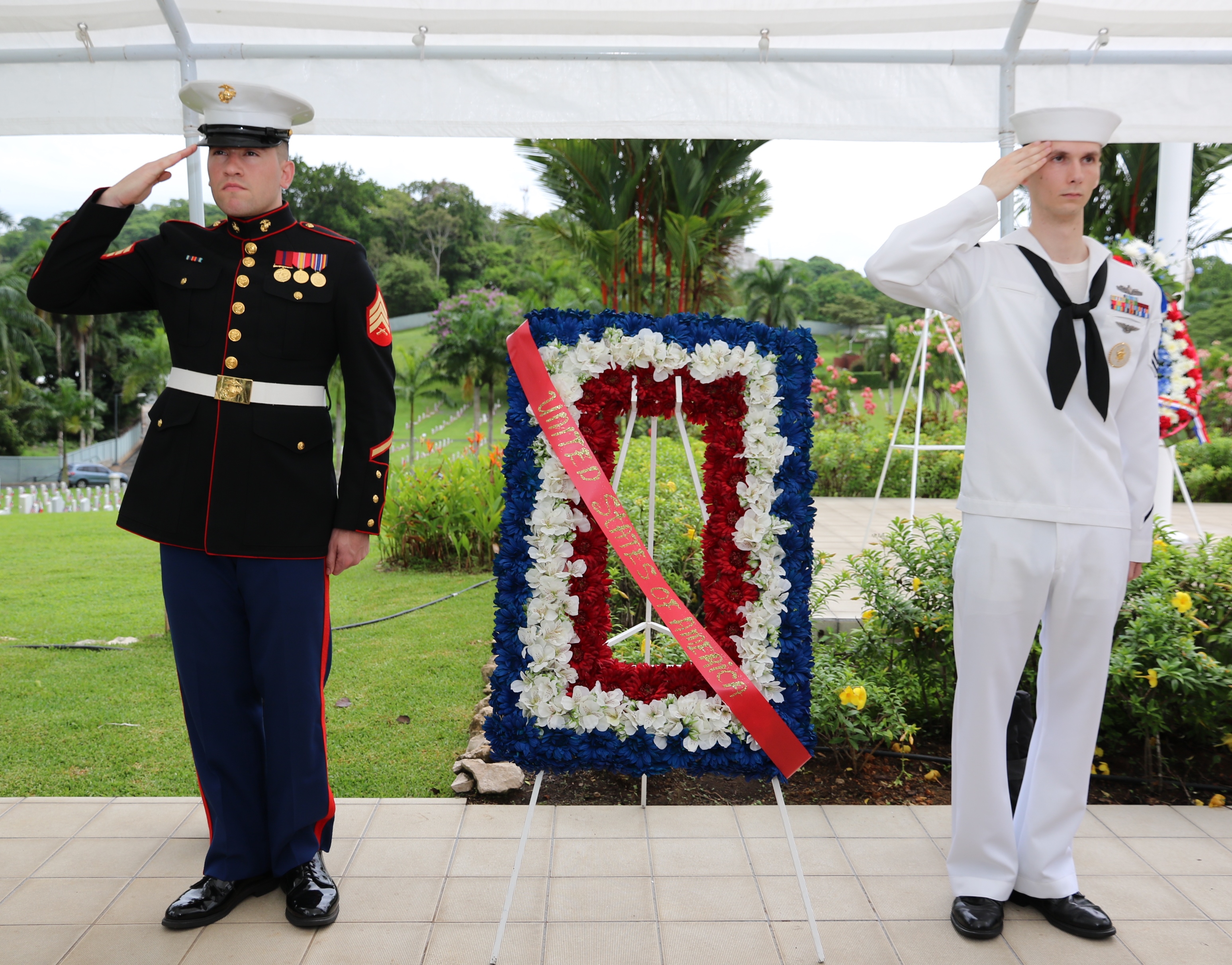 A marine and a sailor salute next to a floral wreath. 