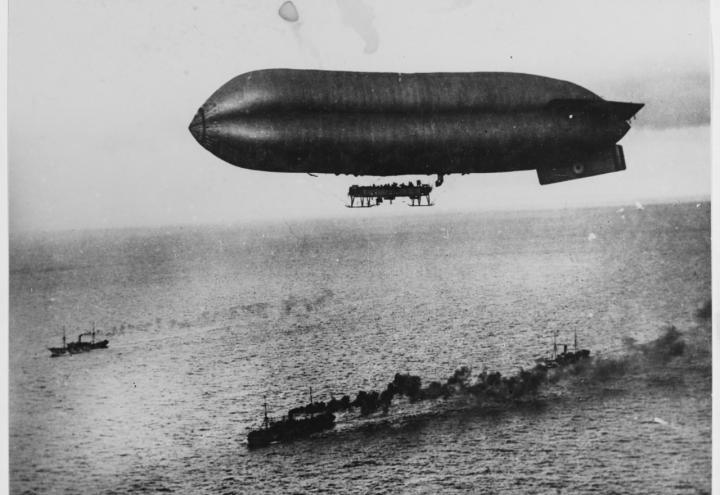 Historic photo showing an airship that looks like blimp flying over large Allied ships. 
