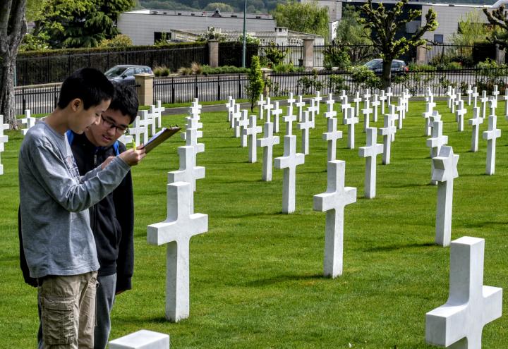 Two students review a paper while standing amongst the headstones. The Eiffel Tower can be seen in the background. 