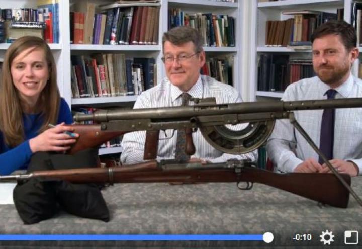 Three people sit at table with large firearms on table in front. 