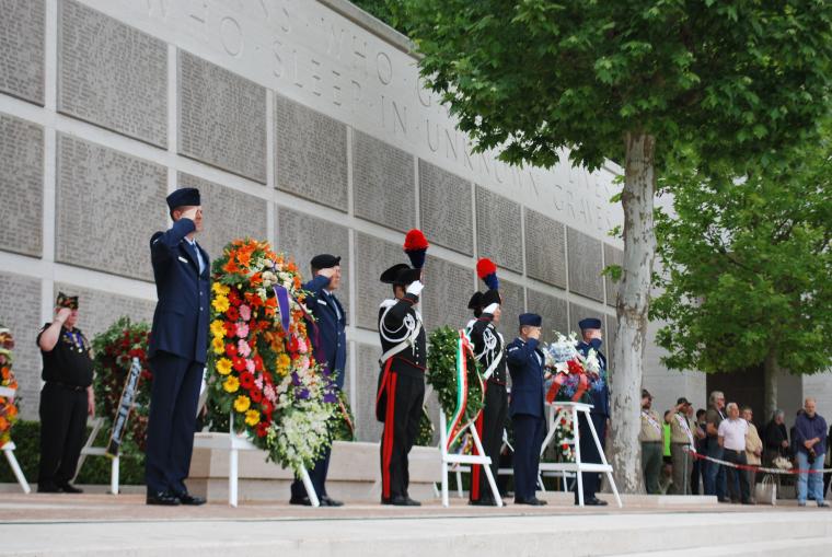 Members of military salute next to floral wreaths. 