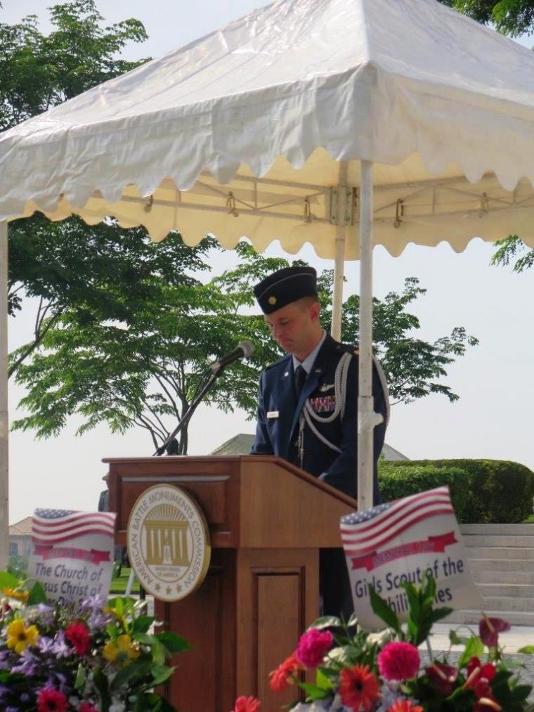 Maj. Rasinski stands at a podium, under a small tent, delivering remarks.