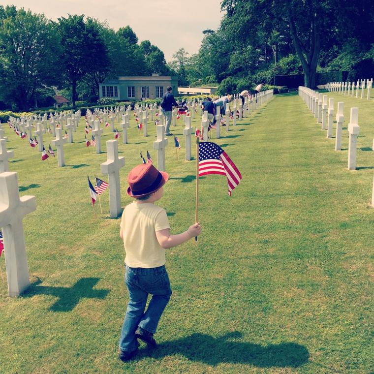 A little boy in a hat carries the American flag as he walks amongst the headstones. 