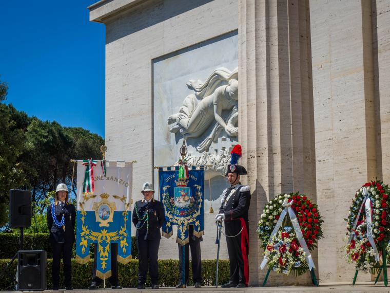 Men and women in uniform stand next to the city banners for Anzio and Nettuno.