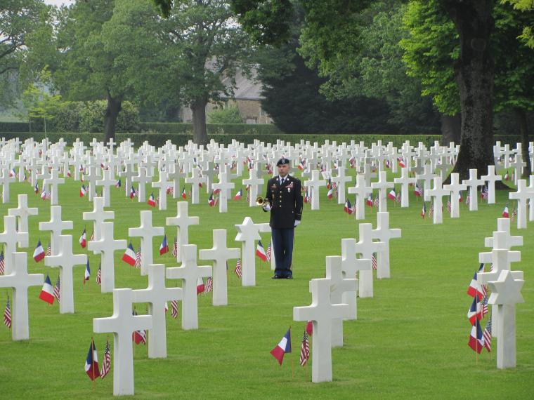 A man in uniform stands amongst the headstones with his trumpet in hand. 