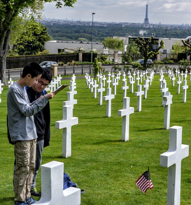 Two students review a paper while standing amongst the headstones. The Eiffel Tower can be seen in the background. 