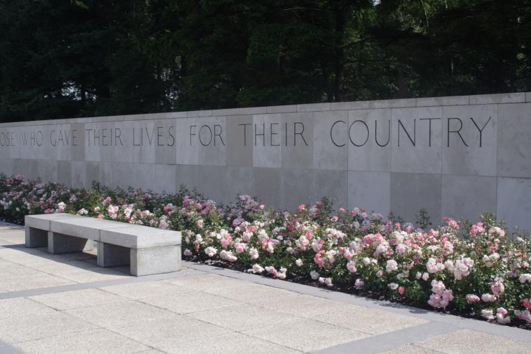 The stone wall includes an inscription, only part of which can be read: "…who gave their lives for their country." Flowers fill the space at the bottom of the wall.