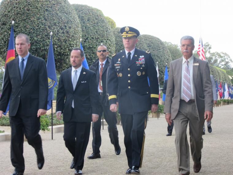 Four men in suits and one in uniform walk along a gravel path of the cemetery.