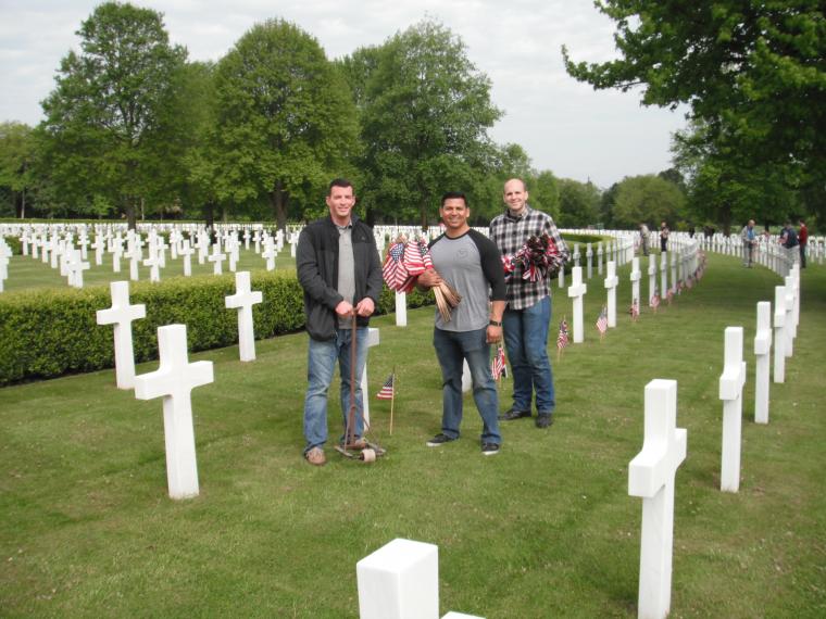 Three men stand with American and British flags amidst the headstones.