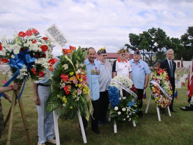 Men stand next to the floral wreaths on stands. 