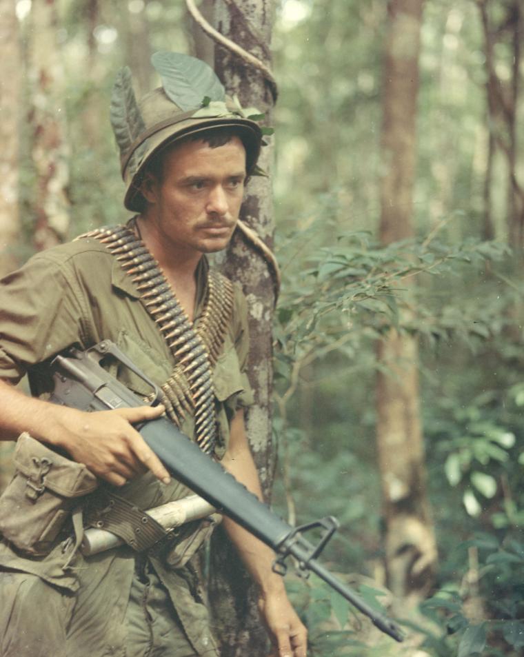 A soldier, wearing jungle-green colored fatigues, leans against a tree with weapon in hand. 
