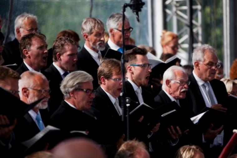Members of the male choir, all wearing dark suits, stand and sing.